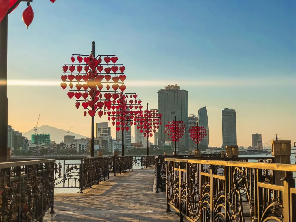 A view of a narrow pier with guardrails, with whimsical heart ornaments on poles lining one side, and skyscrapers in the distance