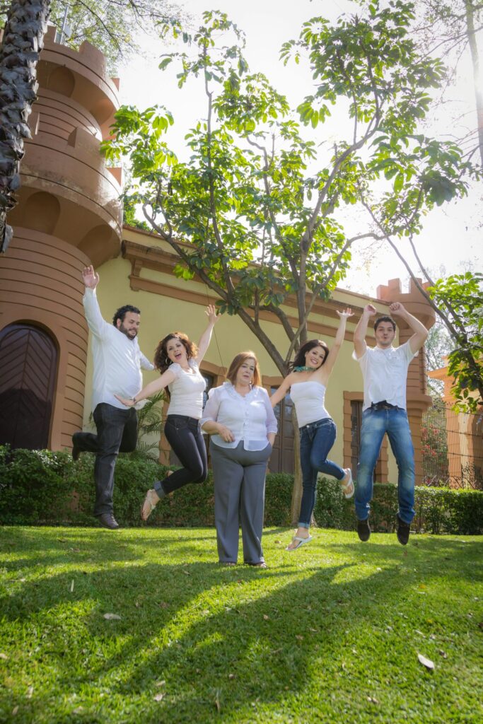 four younger people jump, while a fifth middle-aged woman stands in the middle, in front of a house and tree.