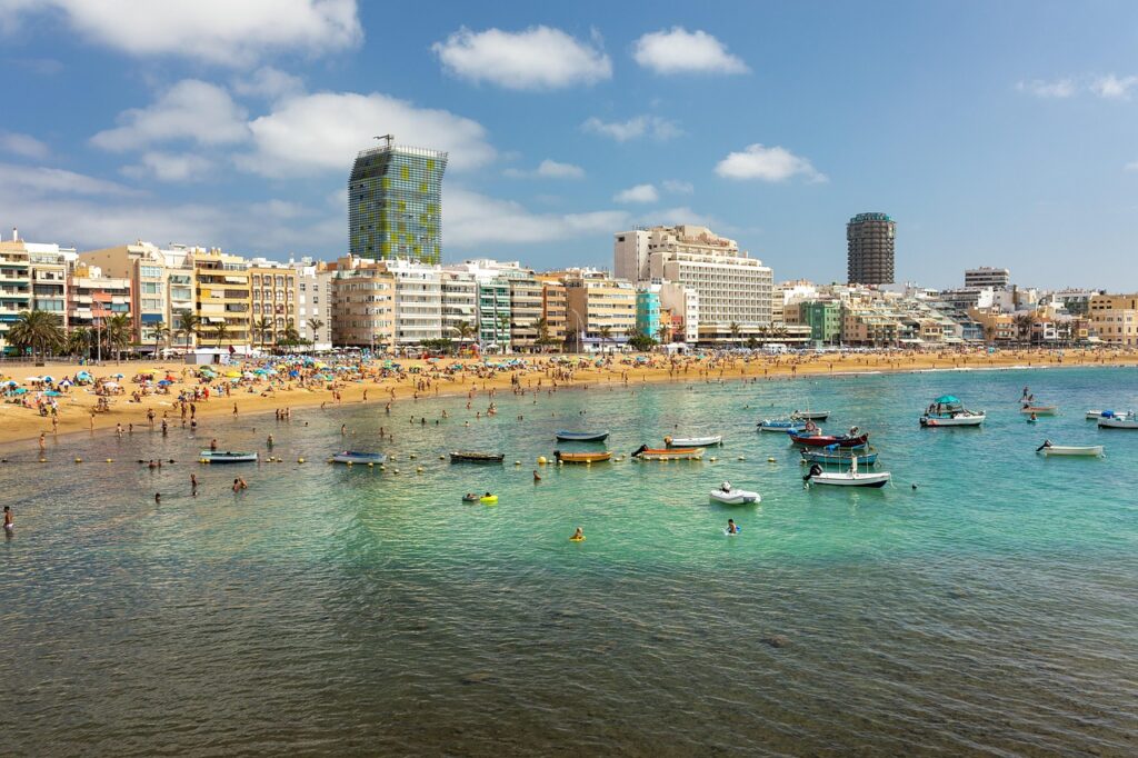Viewed from above the water, a beach filled with people, flanked by 6 story buildings and a few skyscrapers in the background, and numerous boats in the foreground