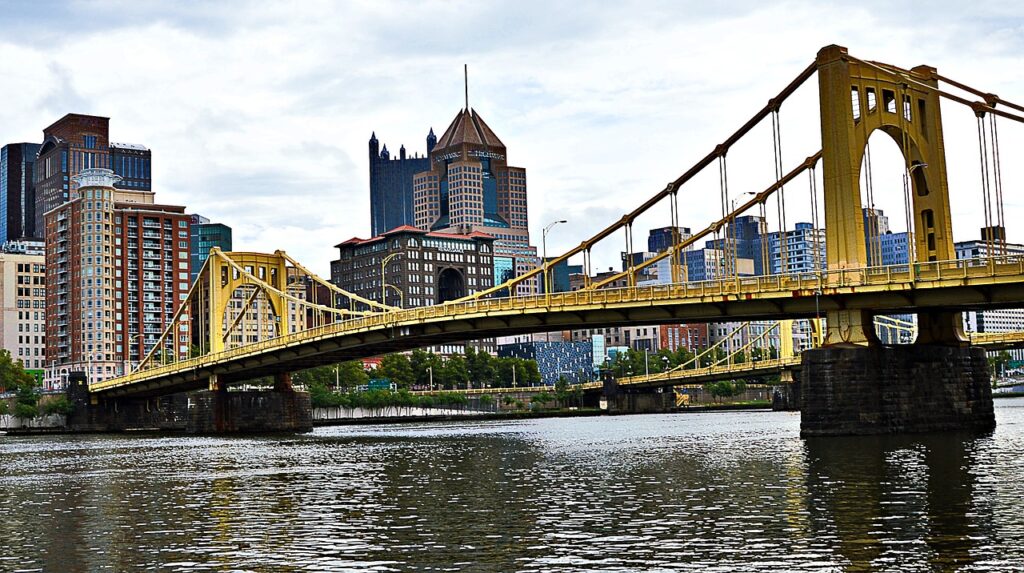 A yellow suspension bridge crosses a river, with skyscrapers on the other side