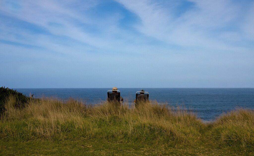 Two people sit in beach chairs in the distance, facing an open sea from grassy dunes.