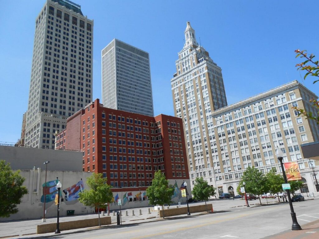 Viewed from a sidewalk, a collection of skyscrapers behind a concrete plaza and a few trees