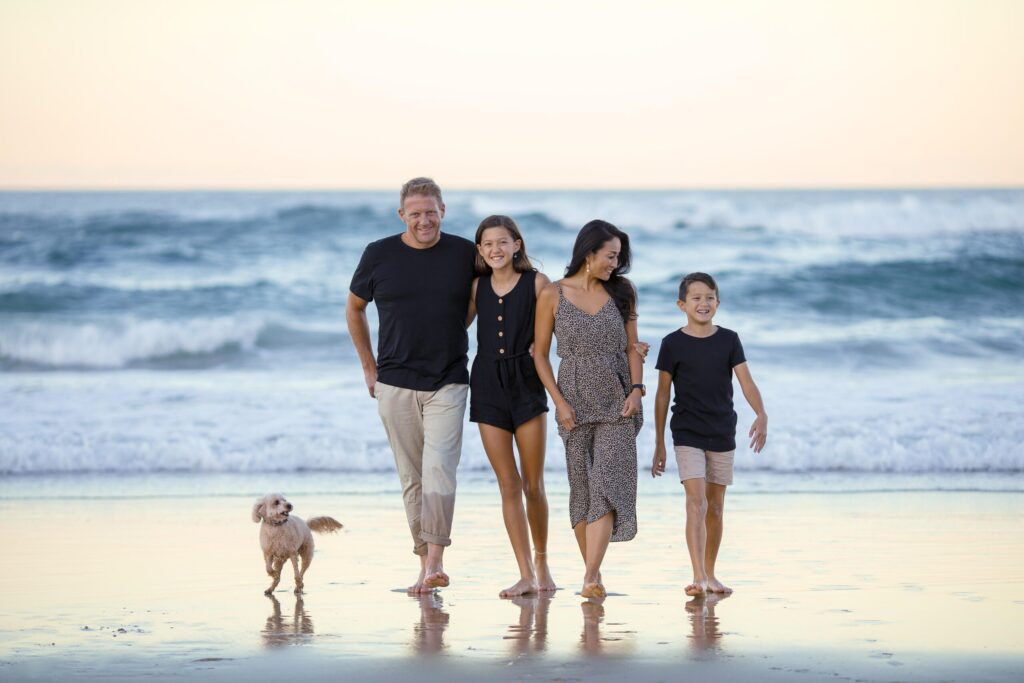 A family of light-skinned people walks towards the camera on the beach, with a wavy ocean behind them.
